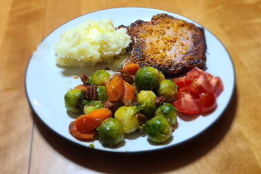 Roasted carrots and brussels sprouts plated with pork, potatoes, and tomatoes.